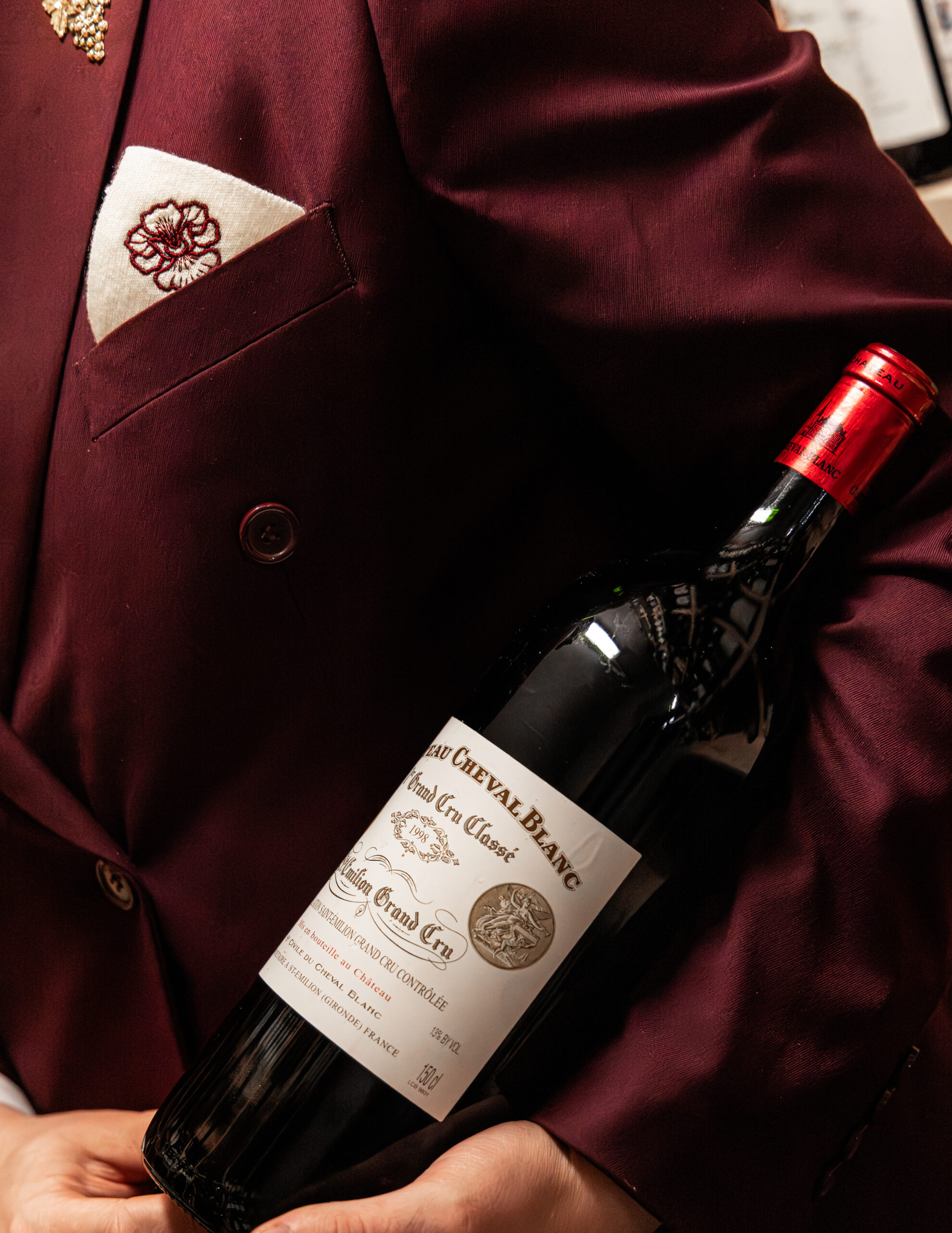 Détail d’une veste bordeaux brodée tenant une bouteille de vin.